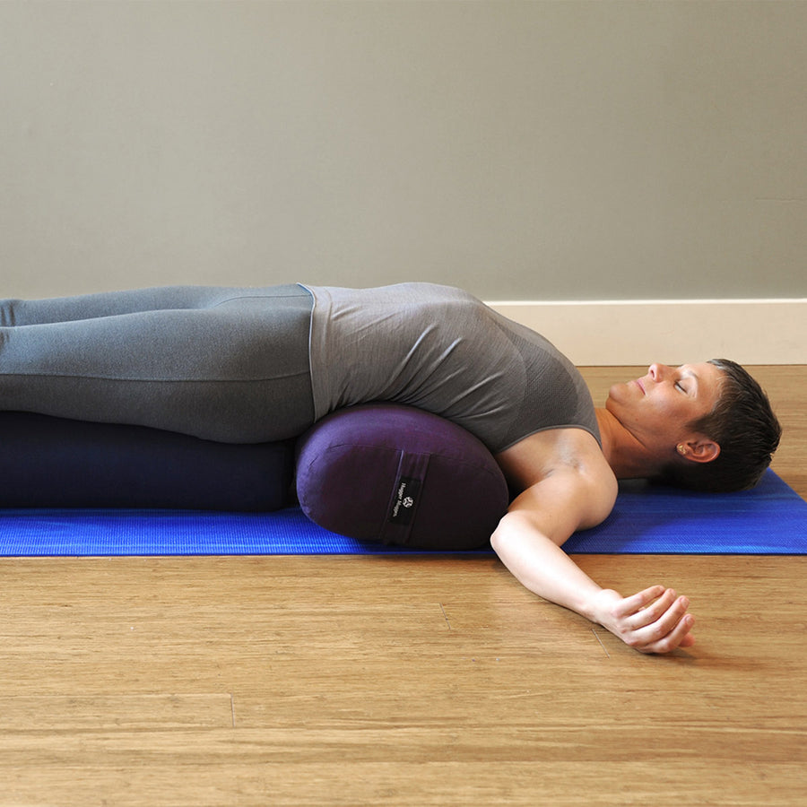 Person lying face up on a mat with the torso supported along a plum yoga bolster