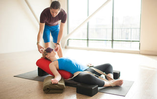 Woman in Supta Baddha Konasana with Standard Bolster - Poppy, Recycled Foam Blocks - Black, D-Ring Strap - Natural, Peachskin Eye Pillow - Plum, Wool Blanket - Olive