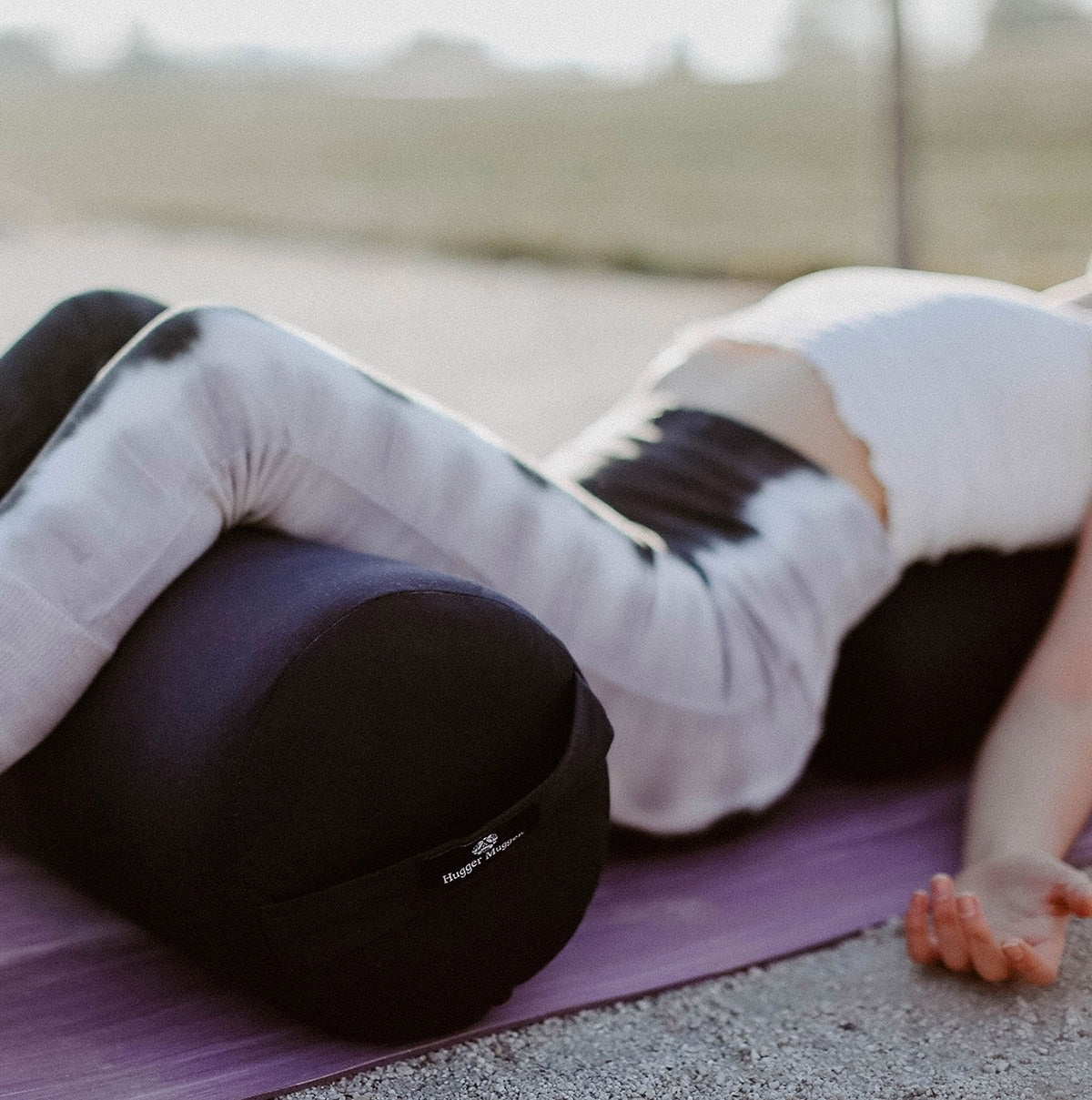 A person is practicing yoga on a mat, using a black bolster for support, with a serene outdoor background.